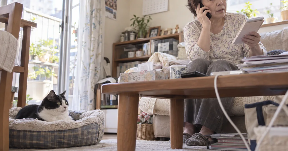 リビングで電話をしながらメモを見る60代の女性と、ペットベッドから見上げる白黒の猫